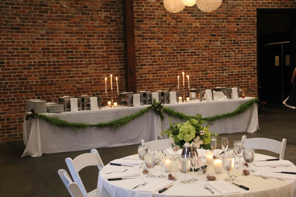 A round table with white linens, floral centerpiece, and glassware is set in front of a buffet table with candles and greenery, against a brick wall.