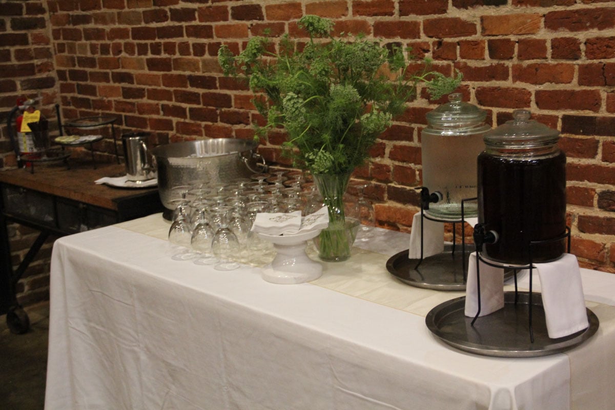 A table with stacked glasses, a large bouquet, and dispensers of water and a dark beverage set against a brick wall.