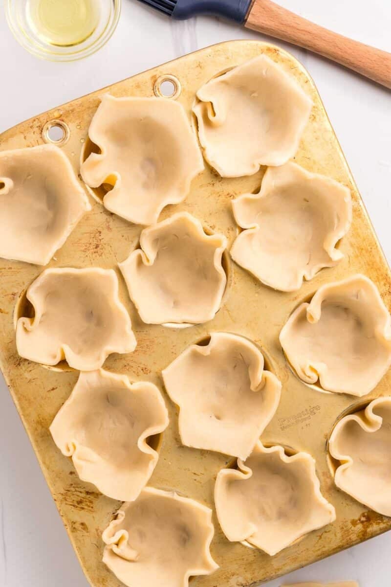 A muffin tin filled with unbaked pie dough pressed into each cup, with a pastry brush and small bowl of oil nearby.