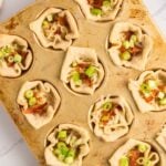 Muffin tin filled with baked pastry cups, topped with shredded cheese, bacon pieces, and chopped green onions, on a light-colored surface.