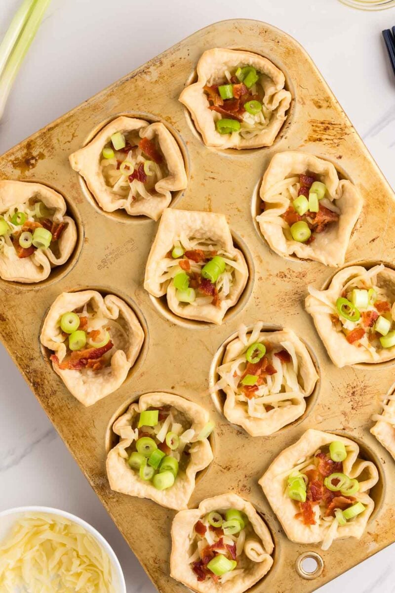 Muffin tin filled with baked pastry cups, topped with shredded cheese, bacon pieces, and chopped green onions, on a light-colored surface.