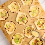 A muffin tin holds twelve mini quiches with chopped green onions on top; a spoon and a bowl of shredded cheese are nearby.