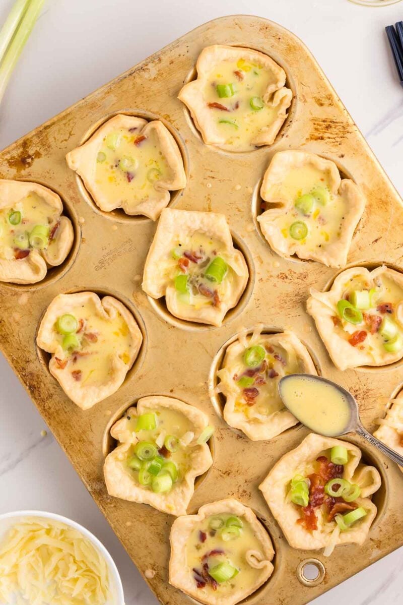 A muffin tin holds twelve mini quiches with chopped green onions on top; a spoon and a bowl of shredded cheese are nearby.