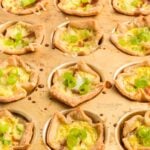 A muffin tin filled with baked mini quiches topped with sliced green onions, each quiche in a pastry crust.