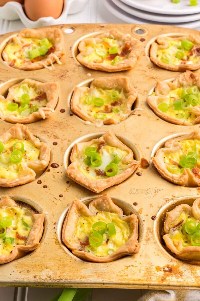 A muffin tin filled with baked mini quiches topped with sliced green onions, each quiche in a pastry crust.
