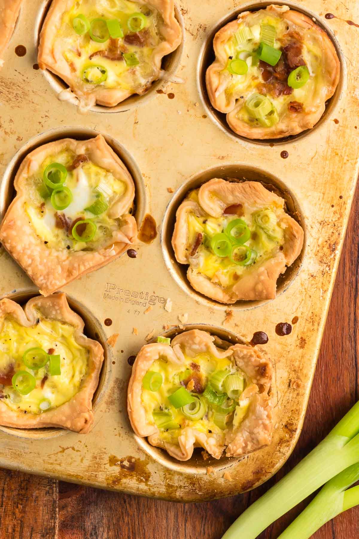 Baked mini quiches with green onions in a muffin tin, on a wooden surface, next to fresh green onions.