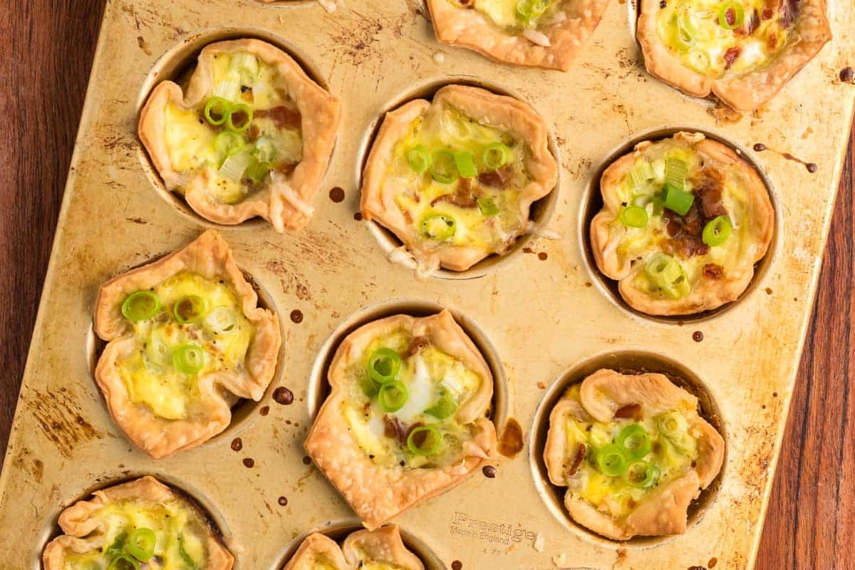 A muffin tin holds several baked mini quiches with crispy edges, filled with egg mixture and topped with chopped green onions.
