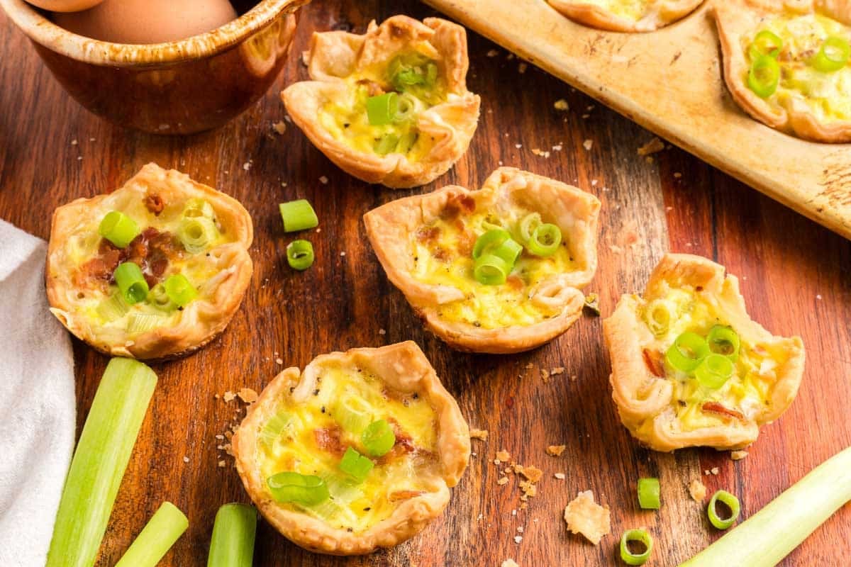 Mini quiches topped with chopped green onions are arranged on a wooden surface, alongside celery stalks, a bowl of eggs, and a baking tray with additional quiches.