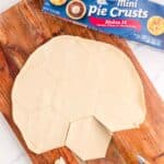 Unbaked pie crust on a wooden board cut into a soccer ball shape, with a knife and scraps nearby; Pillsbury Mini Pie Crusts box in the background.