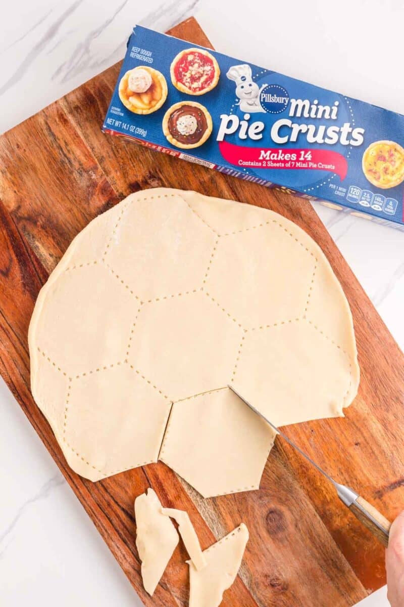 Unbaked pie crust on a wooden board cut into a soccer ball shape, with a knife and scraps nearby; Pillsbury Mini Pie Crusts box in the background.