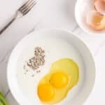 A white bowl with two raw eggs, milk, and black pepper sits on a white surface. Nearby are a fork, a bowl with eggshells, and green onions.
