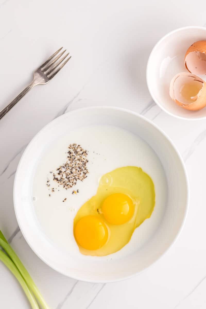 A white bowl with two raw eggs, milk, and black pepper sits on a white surface. Nearby are a fork, a bowl with eggshells, and green onions.