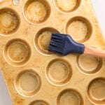 A metal muffin pan with twelve cups is being greased with a silicone pastry brush; some cups have visible oil.