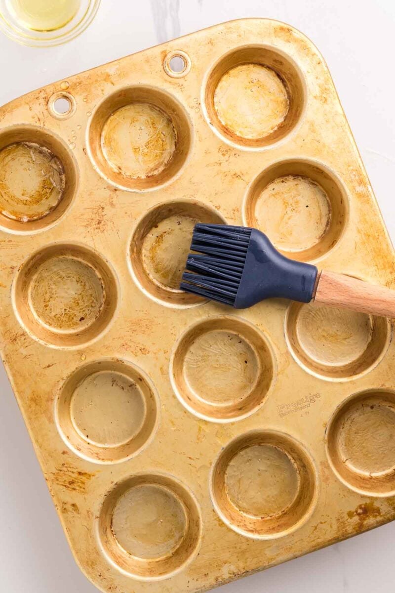 A metal muffin pan with twelve cups is being greased with a silicone pastry brush; some cups have visible oil.
