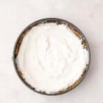 A round baking pan filled with a light, smooth cream mixture on top of a crumbly crust, viewed from above on a neutral surface.