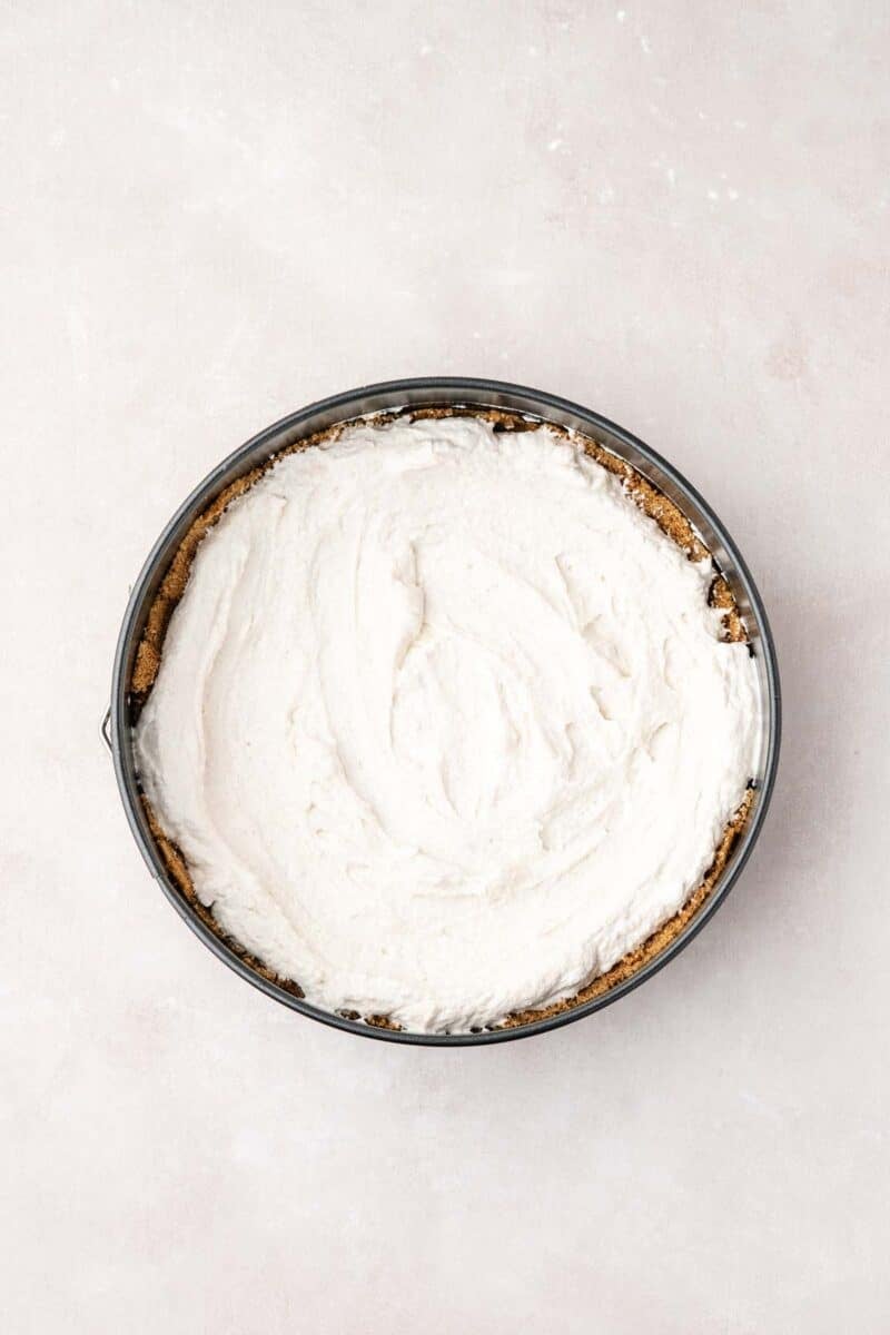 A round baking pan filled with a light, smooth cream mixture on top of a crumbly crust, viewed from above on a neutral surface.