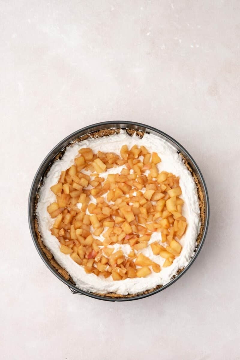 A round springform pan filled with a cream layer, topped with small diced cooked apples, sits on a light surface.