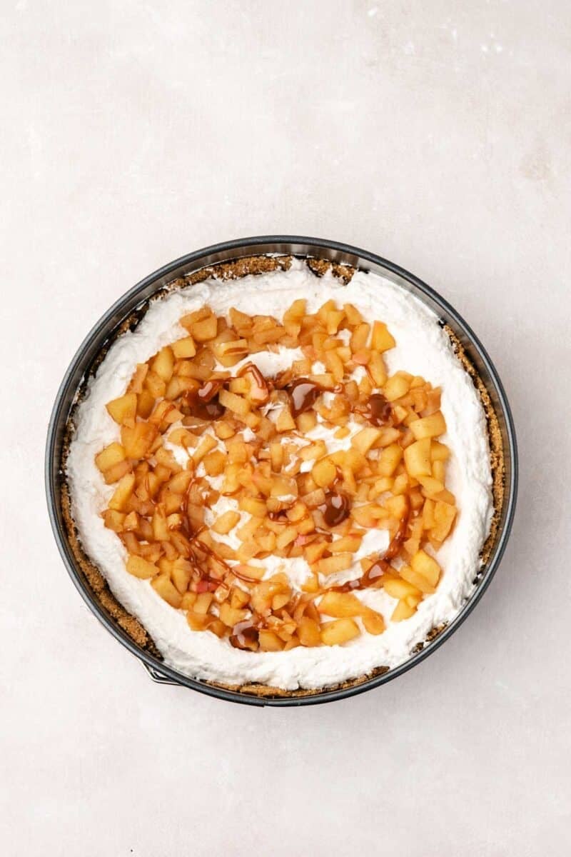 A round pan filled with a cheesecake topped with a swirl of diced caramelized apples and sauce on a light surface.