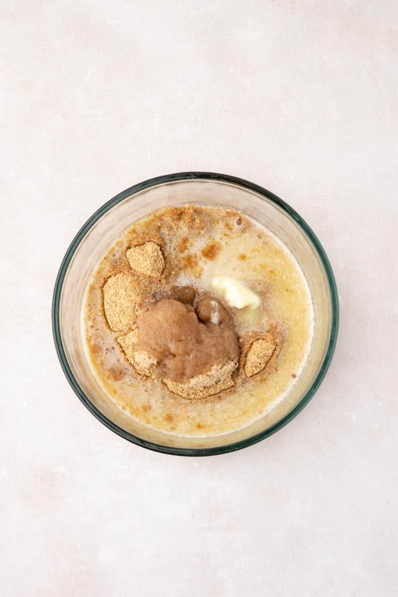A glass bowl containing melted butter, brown sugar, and graham cracker crumbs on a light surface.