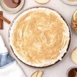 A round apple cheesecake with a crumb topping is shown on a table with apple slices, caramel sauce, cinnamon sticks, and a striped towel.