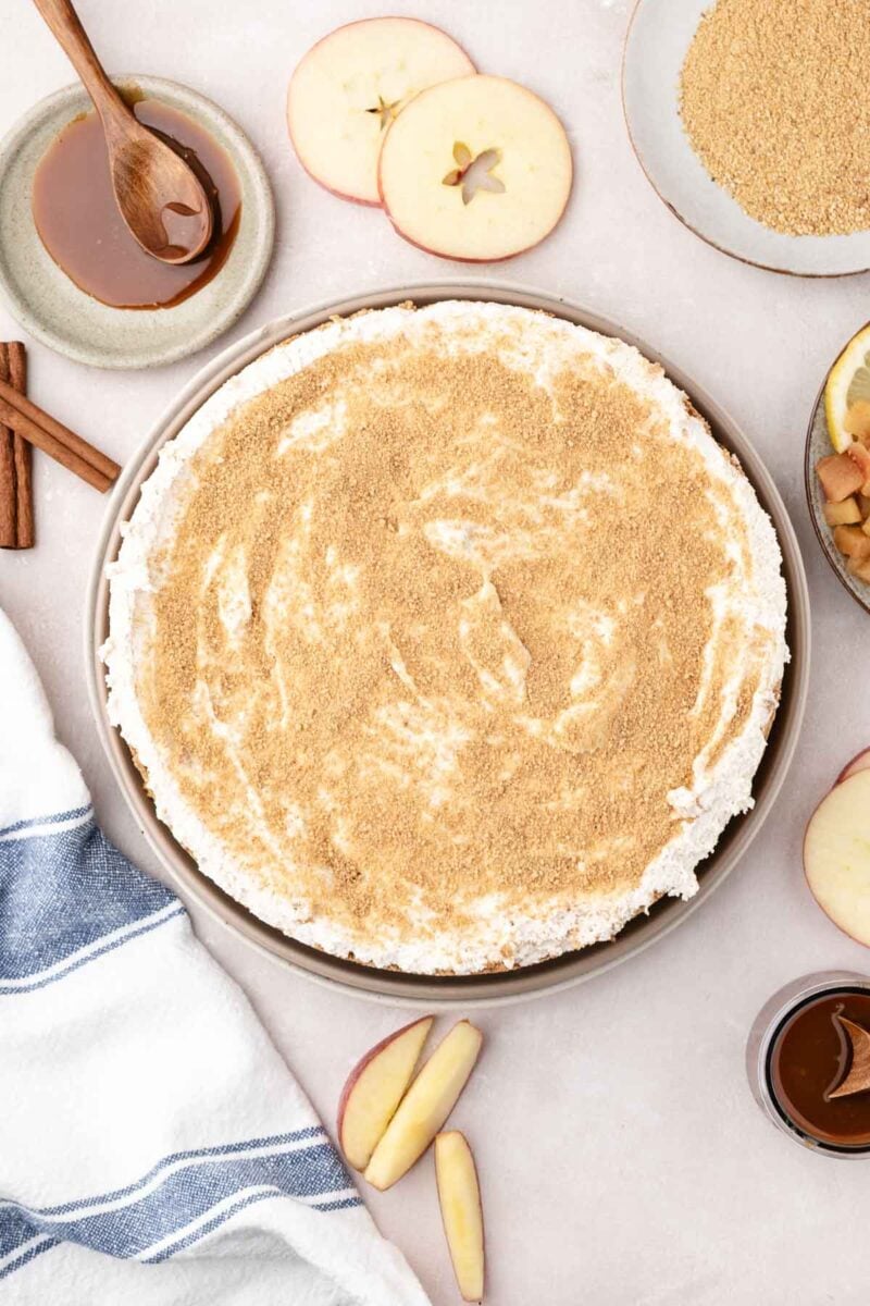 A round apple cheesecake with a crumb topping is shown on a table with apple slices, caramel sauce, cinnamon sticks, and a striped towel.