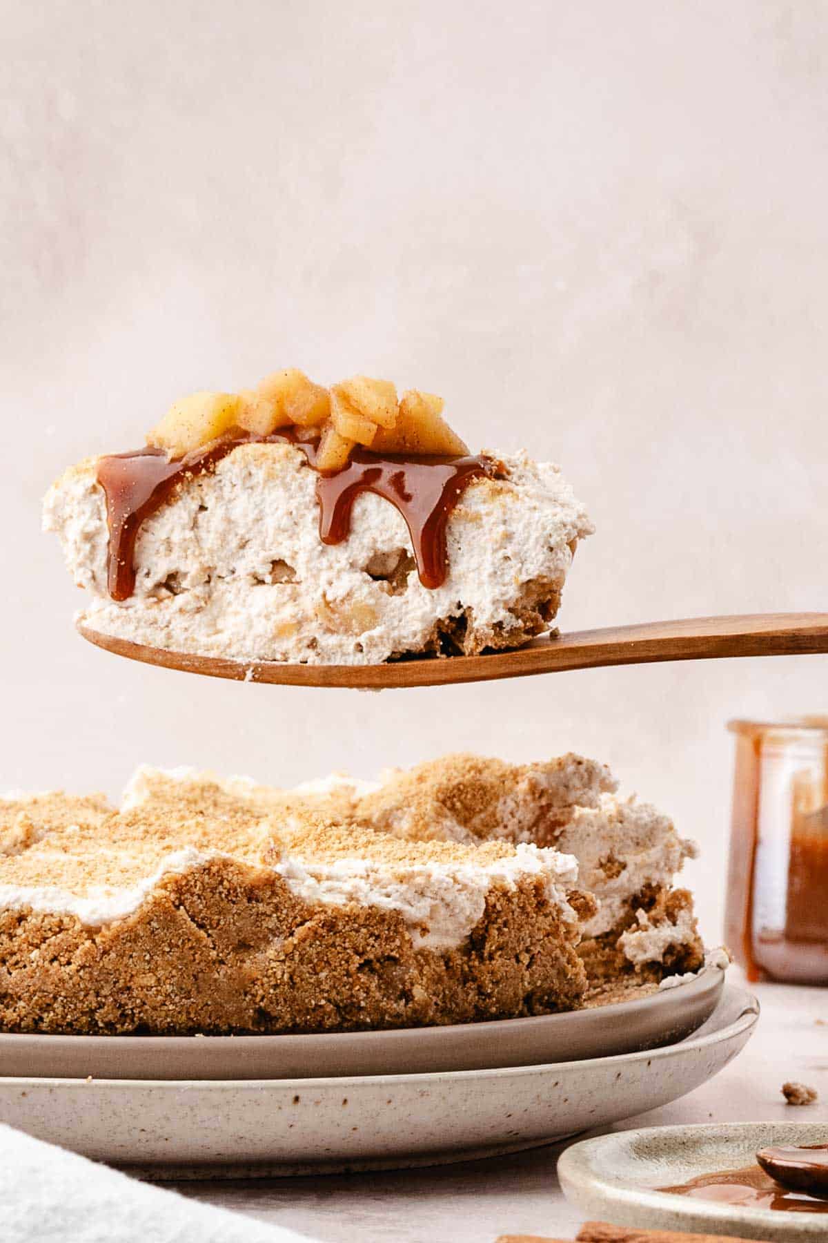 A slice of apple cheesecake with caramel sauce and diced apples is being lifted from a whole pie on a plate.