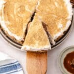 A slice of apple cheesecake with whipped topping is being served from a pie dish, surrounded by caramel sauce and diced apples.