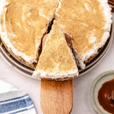 A slice of apple cheesecake with whipped topping is being served from a pie dish, surrounded by caramel sauce and diced apples.