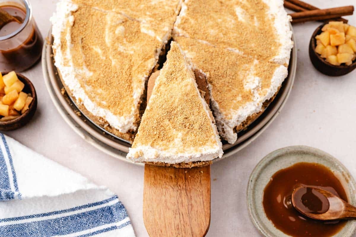 A slice of apple cheesecake with whipped topping is being served from a pie dish, surrounded by caramel sauce and diced apples.