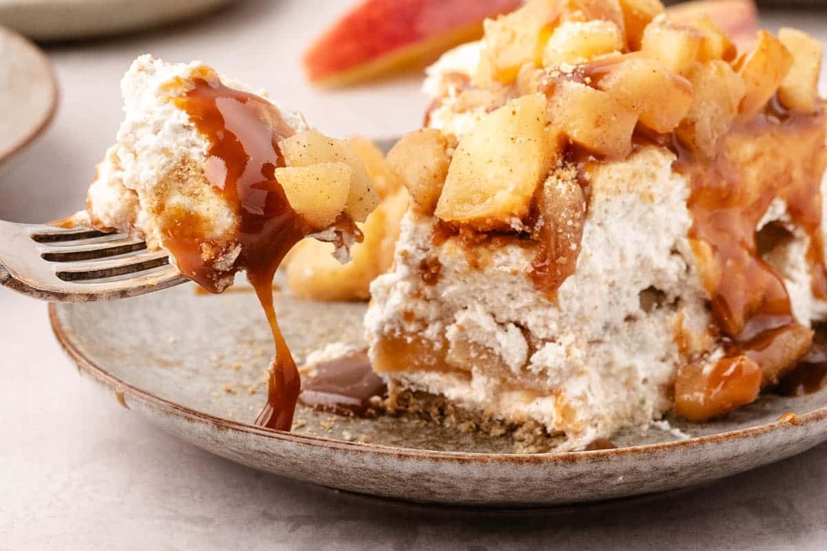 A fork lifts a bite of apple cheesecake topped with caramel sauce and diced apples from a plate.