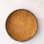 A round springform pan filled with an even layer of pressed graham cracker crust sits on a light-colored surface.