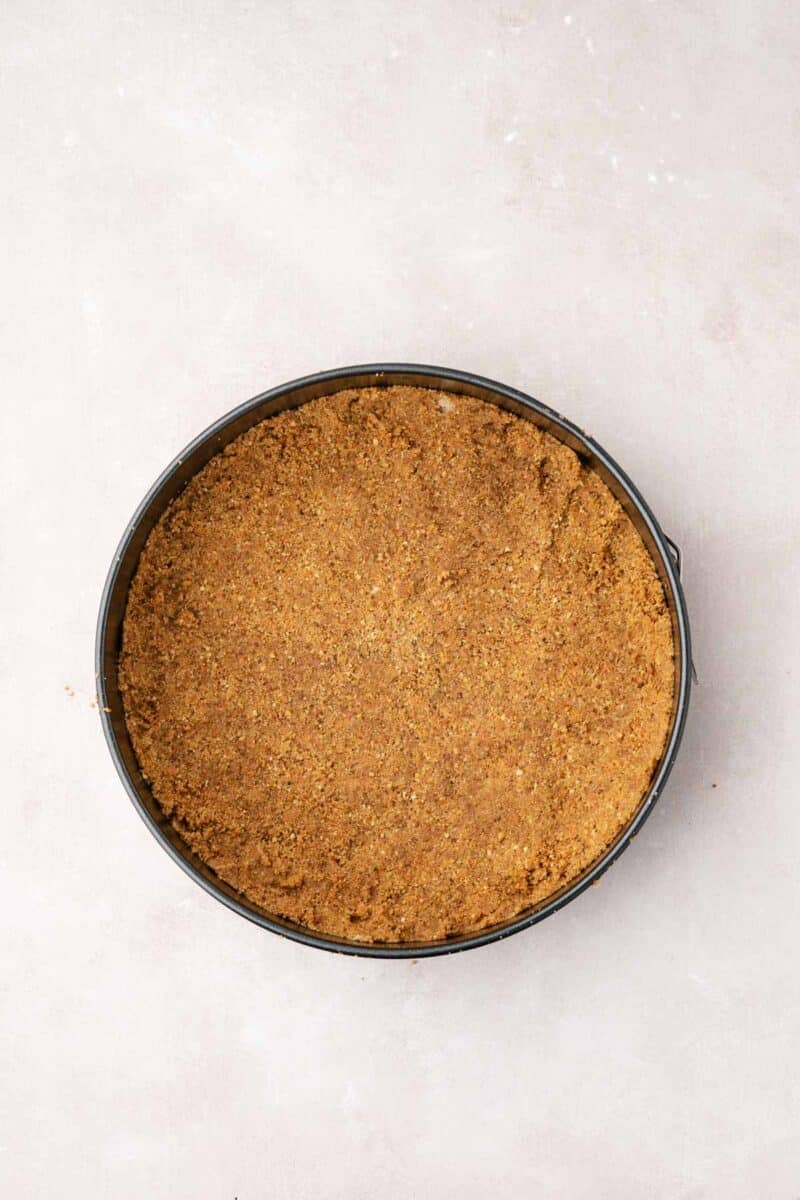 A round springform pan filled with an even layer of pressed graham cracker crust sits on a light-colored surface.