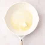 A white frying pan on a light surface contains a partially melted pat of butter.
