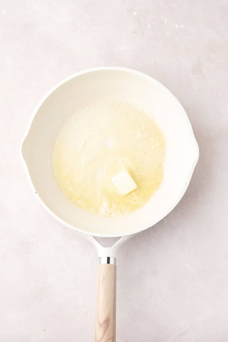 A white frying pan on a light surface contains a partially melted pat of butter.