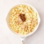 A white frying pan with chopped apples and brown sugar, viewed from above, on a light-colored surface.