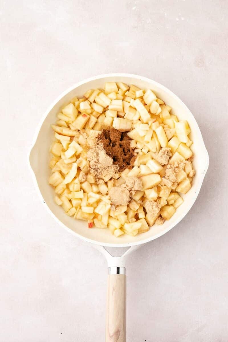 A white frying pan with chopped apples and brown sugar, viewed from above, on a light-colored surface.