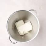Two blocks of cream cheese in a large stainless steel mixing bowl on a light-colored surface.