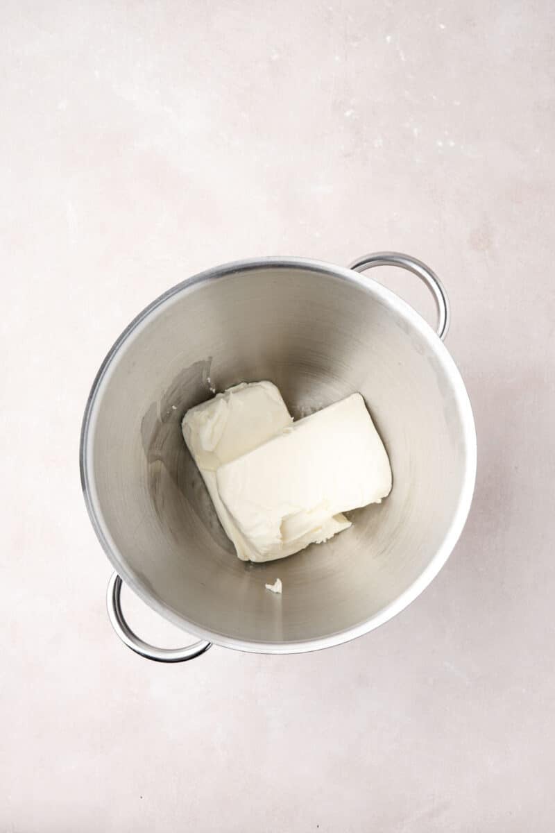 Two blocks of cream cheese in a large stainless steel mixing bowl on a light-colored surface.