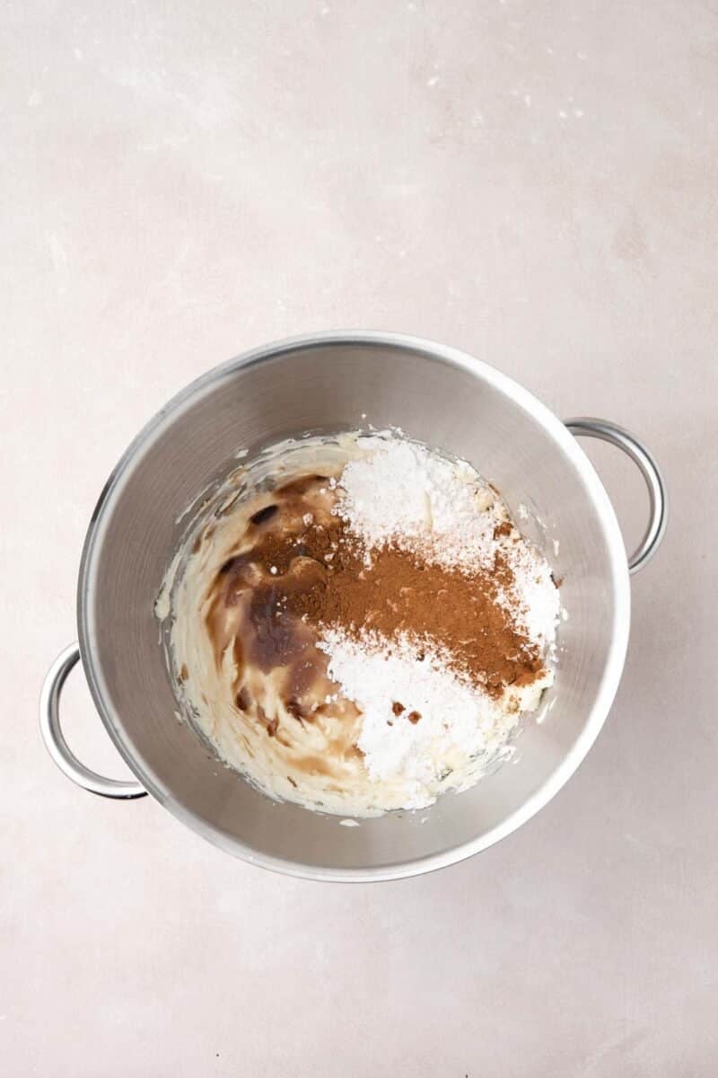 A mixing bowl with brown sugar, powdered sugar, cream cheese, and cinnamon on a light surface, ingredients not yet mixed together.