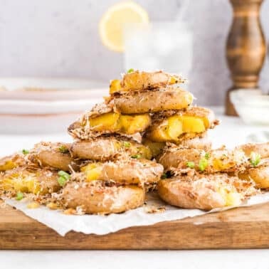 A stack of smashed roasted potatoes topped with grated cheese and chopped herbs sits on a wooden board, with sauce and seasonings in the background.