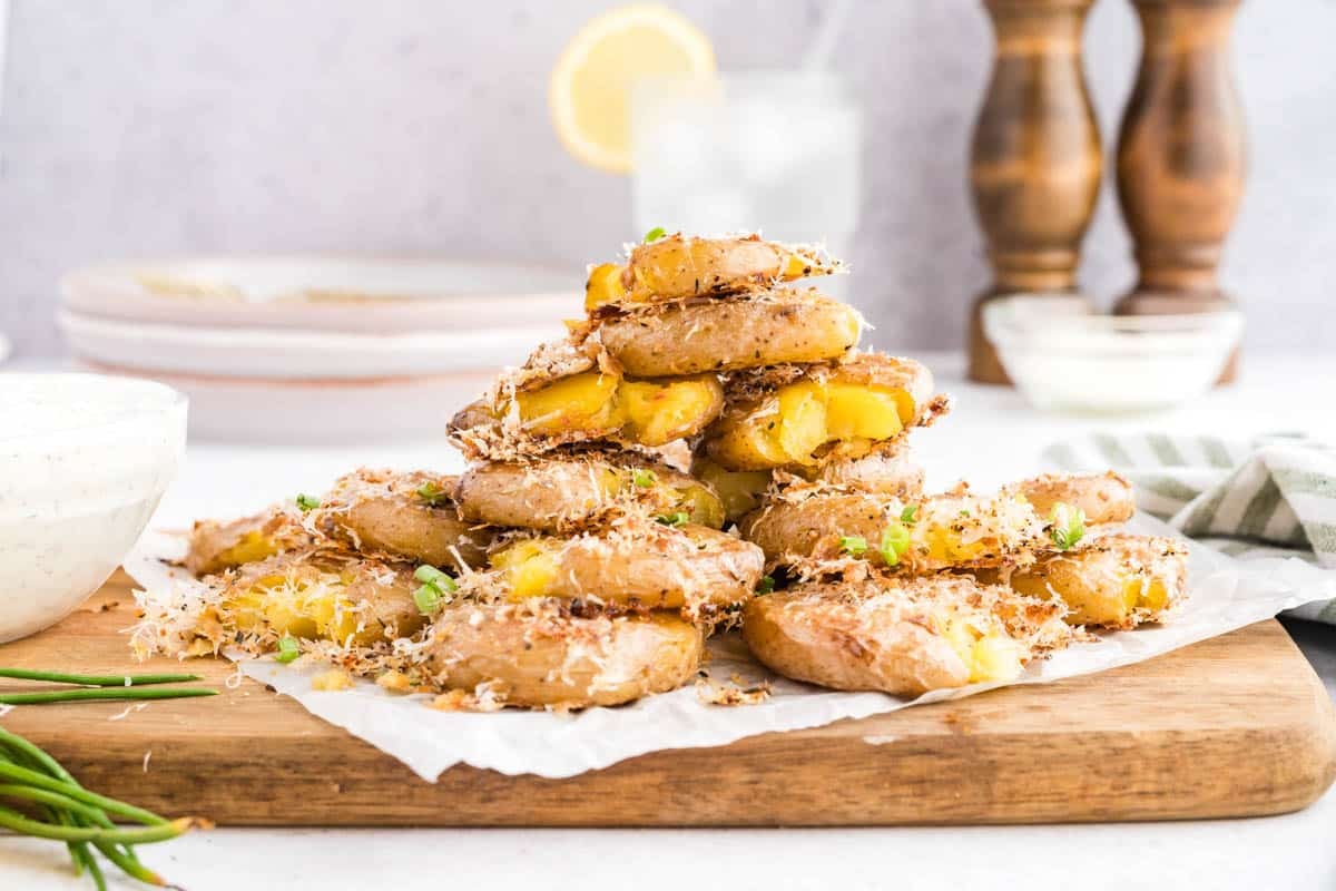 A stack of smashed roasted potatoes topped with grated cheese and chopped herbs sits on a wooden board, with sauce and seasonings in the background.