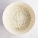 White bowl containing a creamy, herb-speckled sauce or dressing, viewed from above on a light, textured surface.