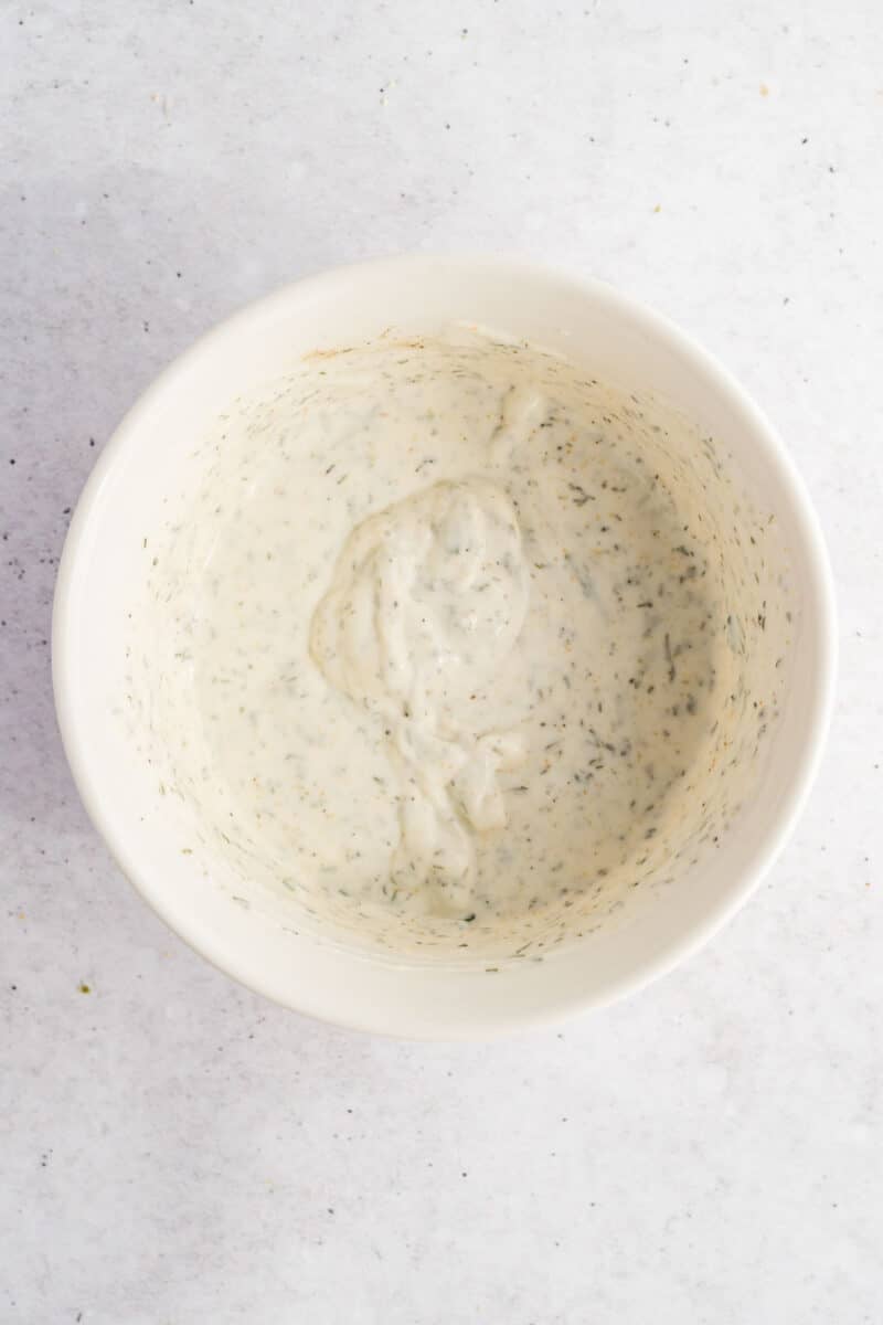 White bowl containing a creamy, herb-speckled sauce or dressing, viewed from above on a light, textured surface.