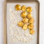 Baked smashed potatoes on a parchment-lined baking sheet, partially covered with shredded cheese, with a bowl of whole potatoes in the background.