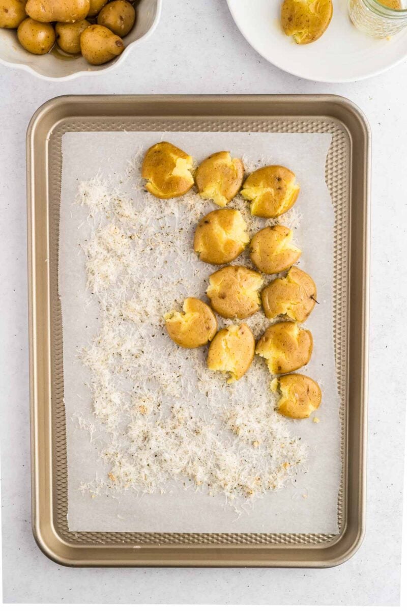 Baked smashed potatoes on a parchment-lined baking sheet, partially covered with shredded cheese, with a bowl of whole potatoes in the background.