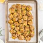 A baking tray of smashed potatoes topped with grated cheese and herbs, set on parchment paper with lemon halves, garlic, and plates in the background.