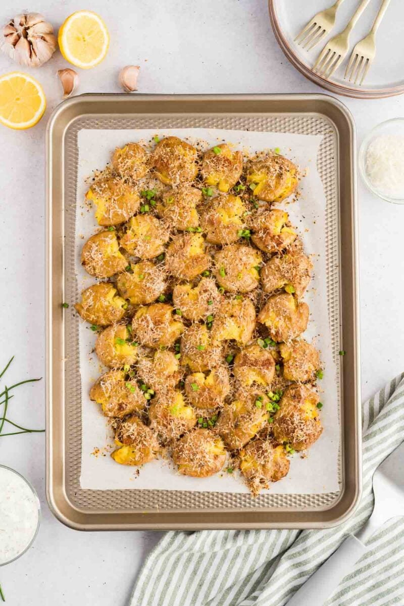 A baking tray of smashed potatoes topped with grated cheese and herbs, set on parchment paper with lemon halves, garlic, and plates in the background.
