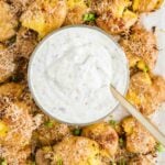 Smashed roasted potatoes topped with grated cheese and chopped green onions, arranged around a bowl of creamy dipping sauce with a spoon.