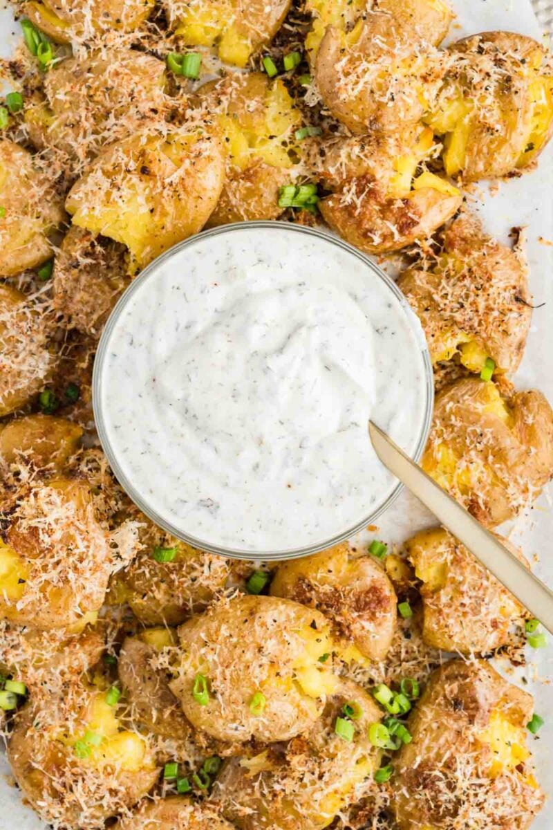 Smashed roasted potatoes topped with grated cheese and chopped green onions, arranged around a bowl of creamy dipping sauce with a spoon.