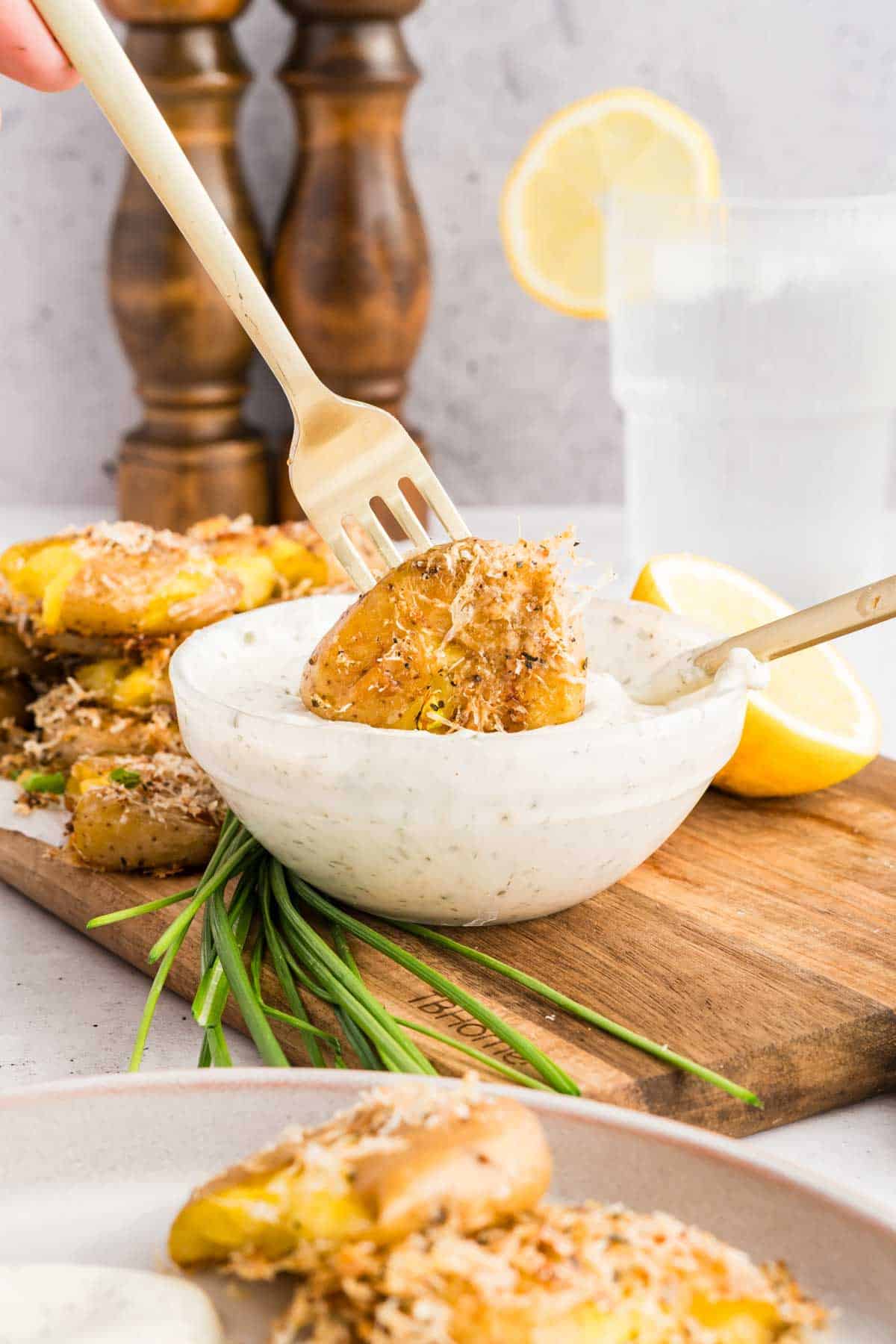 A hand dips a smashed potato with a fork into a bowl of creamy sauce, with lemon, chives, and more potatoes on a wooden board in the background.